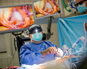 Prof Bakhtiary bei einer minimal-inasiven Herzklappen-OP, bei der er zu Beginn der OP eine 3D-Kamera in den Brustraum und ins Herz schiebt und dann die ganze OP auf den großen Screens im OP-Saal verfolgt. Die beiden Monitore hinter ihm sind die Screens, auf denen der zweite Operateur die OP sehen kann