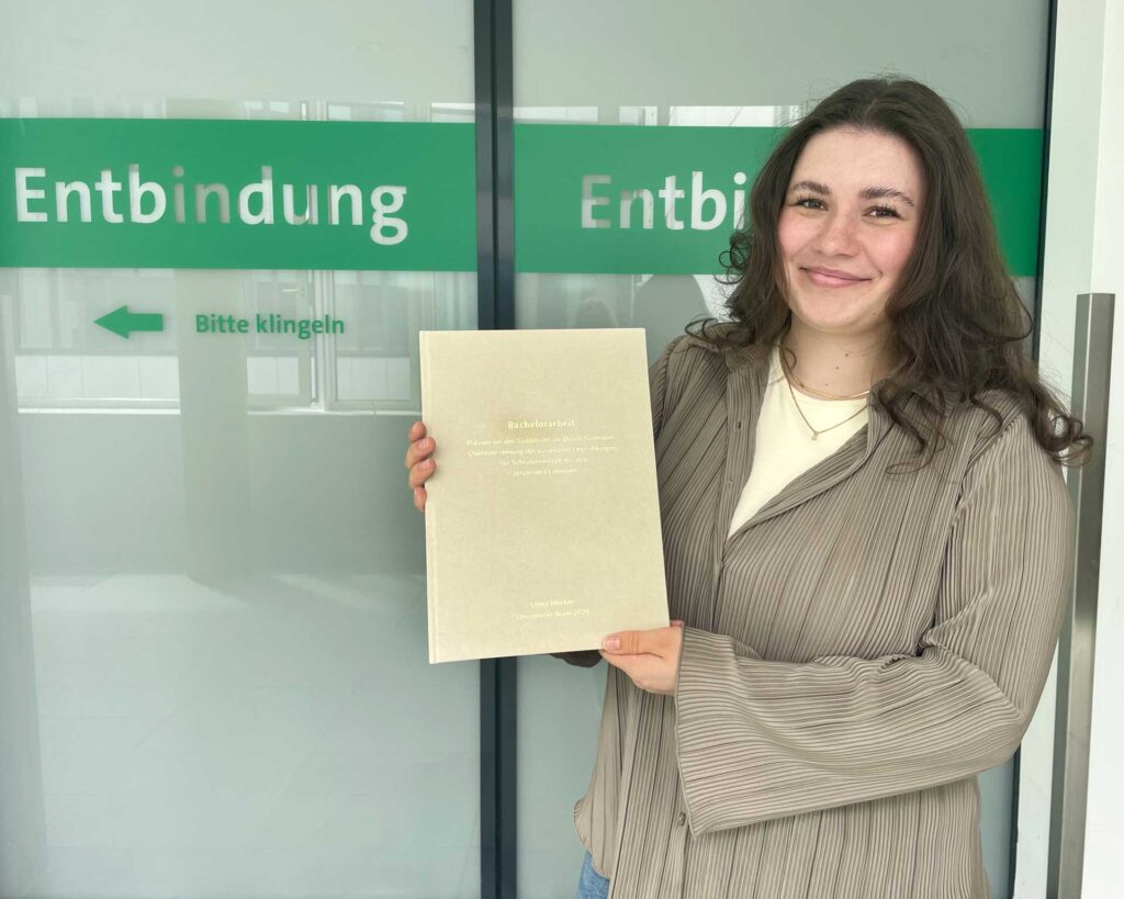 Elena Hecker vor einem Kreißsaal des UKB, in dem sie während ihres Studiums viel Zeit verbracht hat. In den Händen hält sie eine persönliche Druckversion ihrer Arbeit – die offizielle Abgabe erfolgte digital. Foto: Yasmin Kalkan/Medizinische Fakultät Bonn.